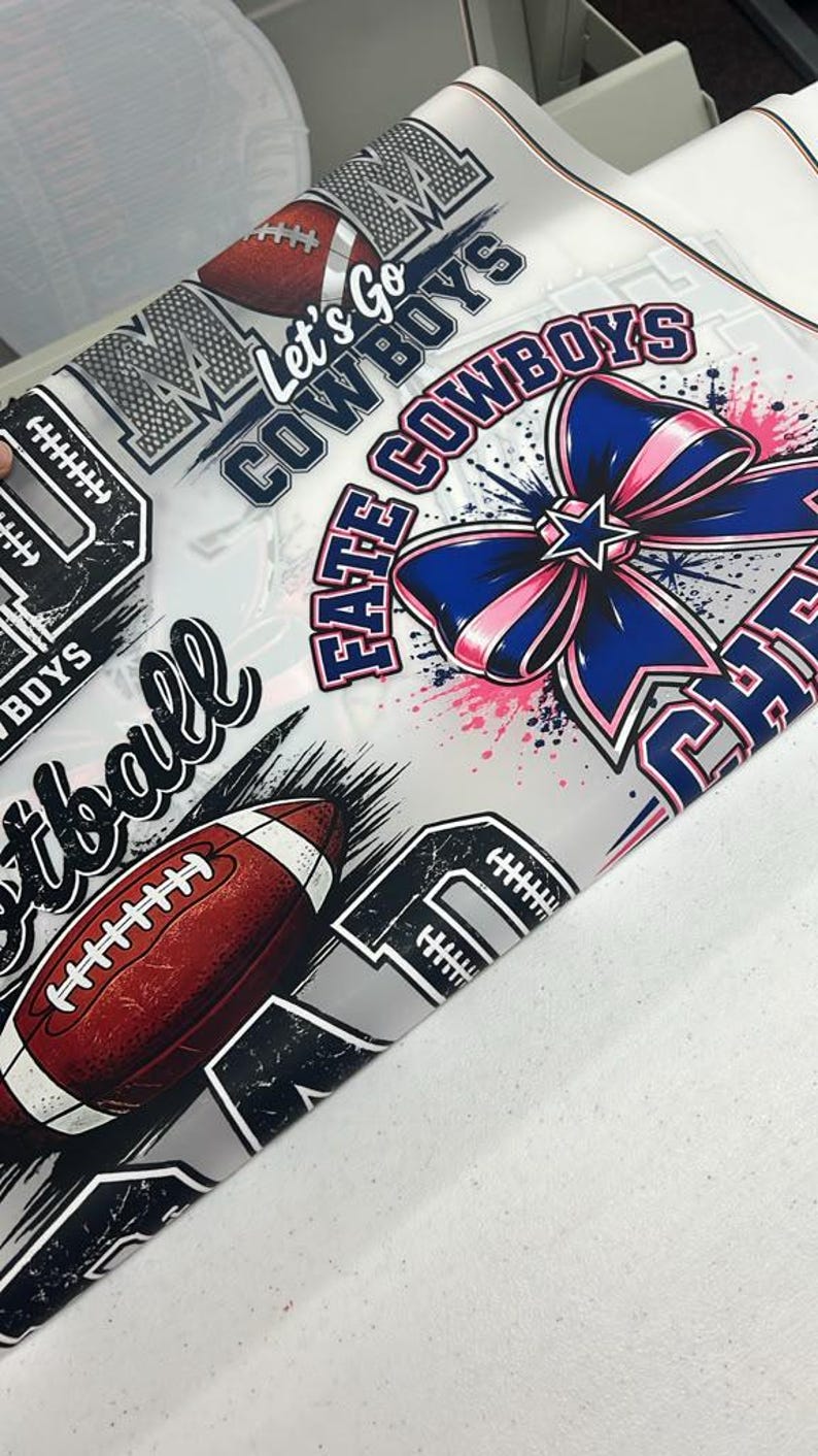 May include: A white fabric sheet featuring a repeating pattern of football-themed graphics, including brown footballs, blue and pink bows, and text that reads Let's Go COWBOYS, FATE COWBOYS, and football. The design includes black and white accents.
