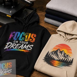 May include: A black hoodie featuring a colorful, distressed-style graphic that reads "FOCUS ON YOUR DREAMS" and a beige hoodie displaying a mountain landscape illustration with a sunset over a lake, accompanied by a matching transparent transfer sheet.