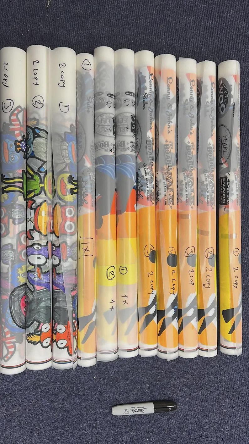 May include: A collection of eleven rolled paper menus or posters, some featuring colorful cartoon illustrations and others displaying the text 'Lenny & John's Boardwalk' and 'The Woo'. A black Sharpie marker is positioned horizontally below the rolls.