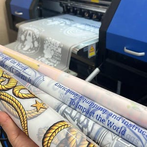 May include: A large format blue and black industrial printer producing printed vinyl rolls. The rolls feature yellow rope patterns, gold stars, and text including "Impact the World," "Dayvid L. Griggs Jr," and "Enriching, Equipping, and Instructing."