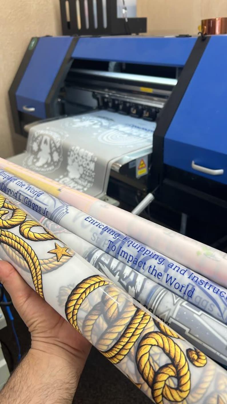 May include: A large format blue and black industrial printer producing printed vinyl rolls. The rolls feature yellow rope patterns, gold stars, and text including "Impact the World," "Dayvid L. Griggs Jr," and "Enriching, Equipping, and Instructing."