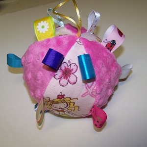 May include: A pink and white fabric ball with various colorful ribbons attached. The ball has a floral pattern and a cartoon girl on one side.