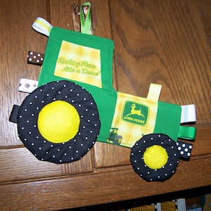 May include: A green and yellow John Deere tractor-themed baby toy with black and white polka dot wheels. The toy has a variety of fabric textures and ribbons for baby to explore. The toy has the text "Nothing Runs Like a Deere!" on the front.