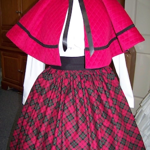 May include: A red quilted cape with black trim, paired with a white blouse and a long red and black plaid skirt. A black ribbon ties the cape. The outfit is a historical costume.