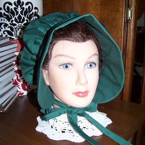 May include: A dark green bonnet with a wide brim and a matching ribbon tie. The bonnet is paired with a white lace collar. The bonnet is displayed on a mannequin head.