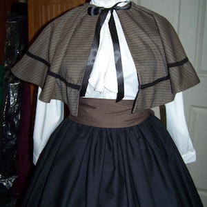 May include: A historical costume featuring a brown and black patterned cape with black trim, a white blouse, and a black skirt. The outfit includes a brown belt and a black bow at the neck, evoking a vintage aesthetic.