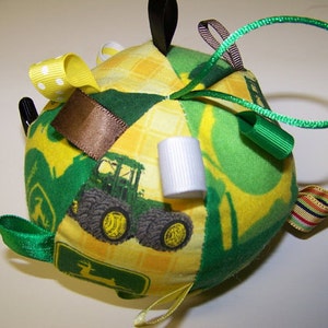 May include: A green and yellow fabric ball with a tractor design and various ribbons attached. The ball is made of different fabrics and textures, including a green and yellow plaid fabric, a green fabric with a tractor design, and a yellow fabric with a deer design.