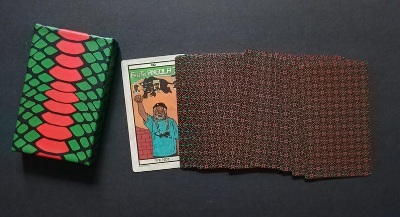 May include: A wallet with a brown and red geometric pattern, a card with an illustration of a person, and a wrapped item with a green and red snake skin pattern. The card has the text "Fist Angola" and "We Not I".
