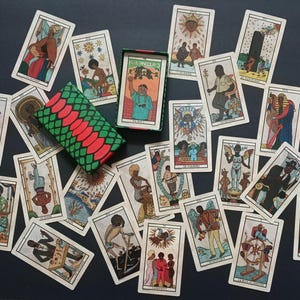 May include: A collection of tarot cards featuring illustrations of people and symbols, spread out on a dark surface. The deck is in a box with a green, red, and black pattern. The cards display various colours and designs.