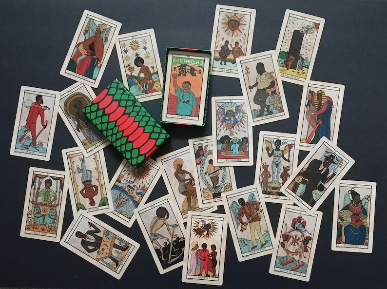 May include: A collection of tarot cards featuring illustrations of people and symbols, spread out on a dark surface. The deck is in a box with a green, red, and black pattern. The cards display various colours and designs.