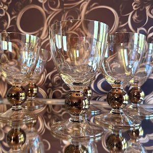 May include: Set of six clear glass goblets with a gold-colored sphere base. The glasses have a classic, elegant design, perfect for serving wine or other beverages. The goblets are arranged on a reflective surface.