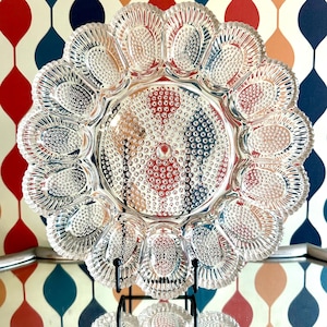 May include: Clear glass deviled egg serving platter with a scalloped edge and a central circular area. The platter has 12 individual egg compartments and is displayed on a black metal stand. The background features a retro-style wallpaper.