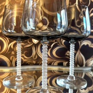 May include: Three elegant wine glasses with a smoky gray bowl and a clear, textured stem. The glasses are arranged on a reflective surface, with a patterned background in shades of gold and brown. The glasses are designed for wine.