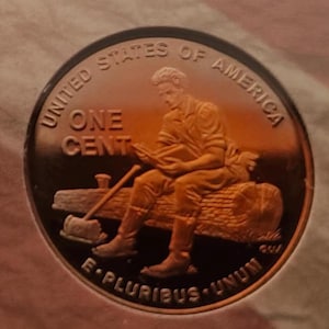 May include: A copper-colored United States one-cent coin. The coin features an image of a man seated on a log, reading. The words "UNITED STATES OF AMERICA", "ONE CENT", and "E PLURIBUS UNUM" are inscribed on the coin.