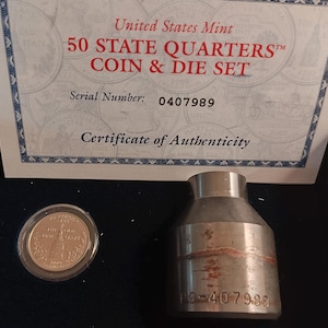 May include: A certificate of authenticity for a 50 State Quarters coin and die set. The certificate is white with blue and gold trim. The serial number is 0407989. The set includes a silver coin and a metal die. The die has the serial number 10-407988 engraved on it.