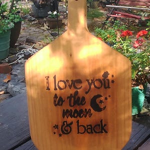 May include: A wooden cutting board with a handle, featuring a burnt-in message that reads "I love you to the moon & back" with a crescent moon and stars.