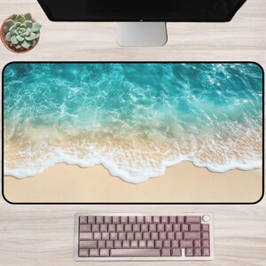 May include: A rectangular desk mat featuring a vibrant beach scene with turquoise water and white waves washing onto a sandy shore. A keyboard with mauve keycaps sits below the mat. A succulent plant in a brown pot is visible.