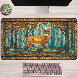 Op de afbeelding: Een rechthoekige bureaumat met een illustratie van een hert in een bos in glas-in-loodstijl. Het kunstwerk gebruikt tinten bruin, goud en blauw. Een toetsenbord staat onderaan en een computermonitor bovenaan.