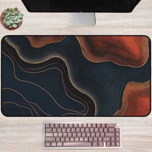 May include: A rectangular desk mat featuring a dark navy blue and burnt orange abstract topographic line pattern. The mat is positioned below a computer monitor and above a compact mechanical keyboard with light purple keys.