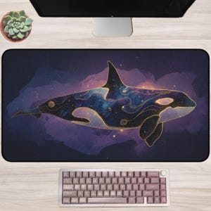 May include: A large rectangular desk mat featuring a stylized orca whale design. The whale is depicted with a cosmic pattern in shades of blue, purple, and gold. The background is a dark, nebula-like design. A keyboard is at the bottom.