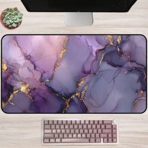 May include: A rectangular desk mat with a purple and gold marble design. The mat has a black border and is next to a keyboard with pink keys. A computer monitor and a small potted succulent are in the background.