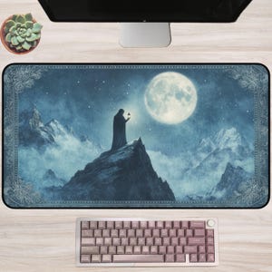 Puede incluir: Una alfombrilla de escritorio rectangular con una ilustración atmosférica en azul oscuro de una figura encapuchada que sostiene una linterna sobre un pico montañoso nevado bajo una gran luna llena, con un borde decorativo.