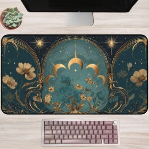 May include: A decorative desk mat featuring a celestial design with gold accents and floral elements. The mat has a teal background with gold crescent moons, stars, and flowers. A mauve keyboard sits below the mat.
