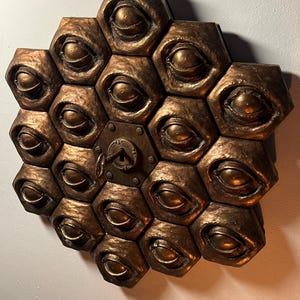 Può includere: Una scultura da parete in metallo dorato con un motivo a nido d'ape di occhi e una serratura al centro.