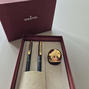 以下が含まれることがあります： マルーンカラーの箱に入ったシェーファーのペンセット。ゴールドトーンのクリップが付いた黒いペン2本と、磨き上げられたゴールドトーンの球体デスクアクセサリーが含まれています。箱の蓋は取り外されており、前面に白いシェーファーのロゴが表示されています。