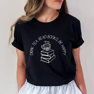 May include: Black t-shirt with a white graphic of a teacup on top of a stack of books. The text "DRINK TEA, READ BOOKS, BE HAPPY" is arched above the graphic. The shirt has short sleeves and a crew neck.