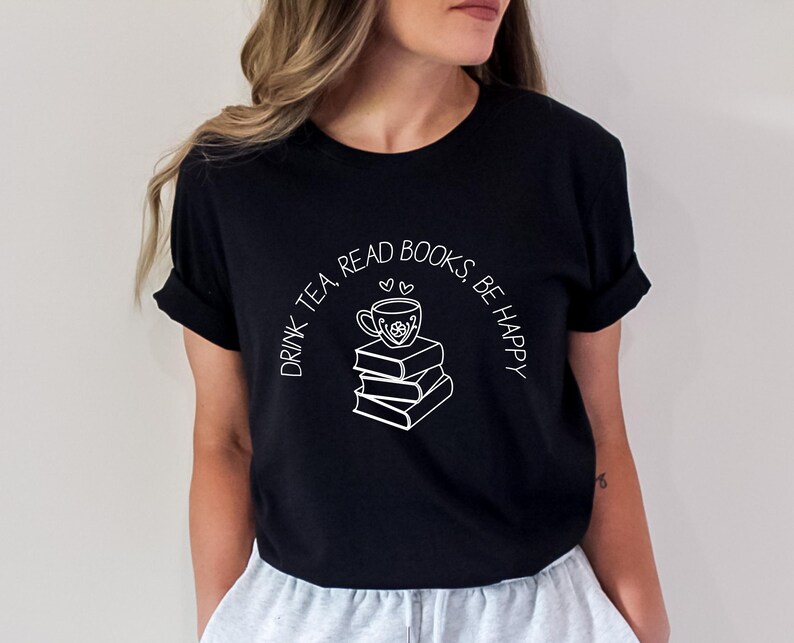 May include: Black t-shirt with a white graphic of a teacup on top of a stack of books. The text "DRINK TEA, READ BOOKS, BE HAPPY" is arched above the graphic. The shirt has short sleeves and a crew neck.