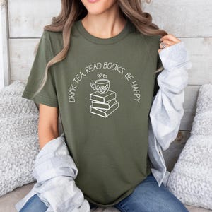 May include: Olive green t-shirt with a white graphic of a teacup on top of a stack of books, with the text "DRINK TEA, READ BOOKS, BE HAPPY" in a curved arc above the graphic. The shirt is made of a soft material.