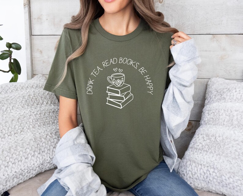 May include: Olive green t-shirt with a white graphic of a teacup on top of a stack of books, with the text "DRINK TEA, READ BOOKS, BE HAPPY" in a curved arc above the graphic. The shirt is made of a soft material.