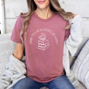 May include: A dusty rose t-shirt with the text "DRINK TEA, READ BOOKS, BE HAPPY" in a curved white font. A white graphic of a teacup over a stack of books is centered on the shirt. The shirt is made of soft material.