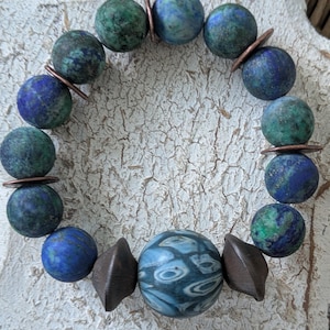 Peut inclure: Un bracelet de perles composé de perles sphériques bleues et vertes, entrecoupées d'entretoises cuivrées. La perle centrale est une sphère bleue plus grande, ornée de motifs, flanquée de perles géométriques marron foncé. Le bracelet est présenté sur une surface texturée blanc cassé.