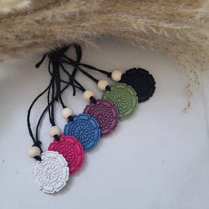 Può includere: Sei profumatori per auto a forma di fiore, in vari colori: bianco, rosa, blu, viola, verde e nero. Ognuno è attaccato a un cordino nero con una perlina di legno. Lo sfondo è bianco con piante essiccate.