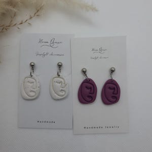 May include: Two pairs of handmade earrings. One pair is white with a face design, the other is a deep purple color with a face design. Each earring hangs from a silver ball stud. The earrings are displayed on white cards with the text "New Brave Jewelry & Accessories".