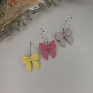 May include: Three pairs of bow-shaped earrings in pastel shades: yellow, pink, and lavender. Each earring has a bow pendant hanging from a silver hoop. The earrings are presented on a white surface, with dried flowers in the background.