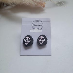 Può includere: Un paio di orecchini a lobo raffiguranti una maschera bianca simile a un fantasma con fori neri per occhi e bocca, incorniciata da un cappuccio nero. Gli orecchini sono esposti su un cartoncino bianco con il testo "Neon Phase Jewelry & Accessories".