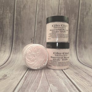 Puede incluir: Tres tarros de manteca corporal Baccarat Rouge de Glo Girl Skincare. Dos tarros están apilados y uno está abierto, revelando un remolino de manteca corporal rosa. Los tarros tienen tapas negras y etiquetas blancas con texto negro. El fondo es un veteado de madera clara.