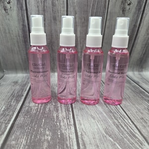 Puede incluir: Cuatro botellas de spray de bruma corporal rosa con tapas blancas están dispuestas en fila. Cada botella tiene una etiqueta con el texto "Glo Girl Skincare" y un nombre de aroma diferente: Pheromones, Cocoa Butter Cashmere, Cheirosa 62 y Baccarat Rouge. Las botellas están sobre una superficie de madera.
