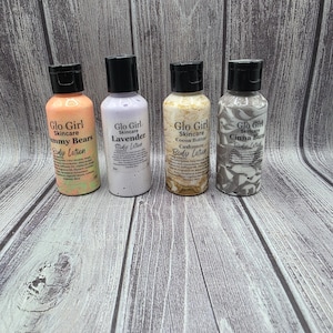 May include: Four bottles of Glo Girl Skincare body lotion are arranged on a wooden surface. The bottles have black caps and labels with the scents: Gummy Bears, Lavender, Cocoa Butter Cashmere, and Cinna Bun. The labels are in orange, purple, beige, and gray.