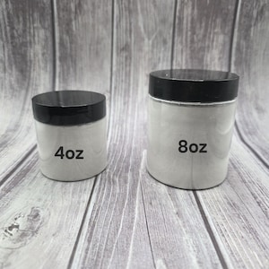 May include: Two white jars with black lids are displayed. The smaller jar is labeled "4oz" and the larger jar is labeled "8oz". The jars are filled with a white substance and sit on a wooden surface.