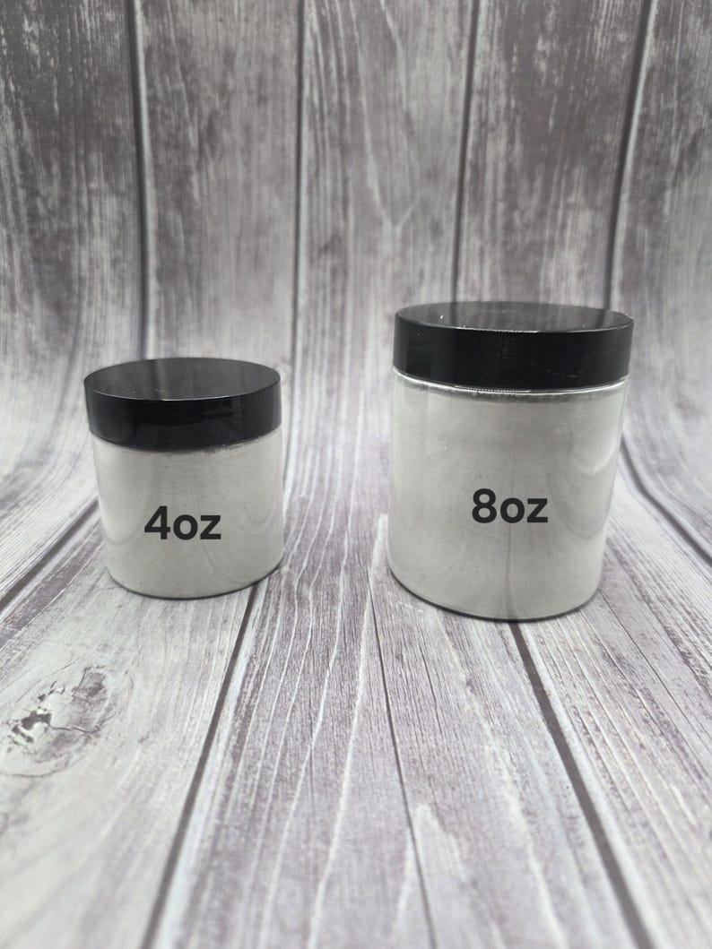 May include: Two white jars with black lids are displayed. The smaller jar is labeled "4oz" and the larger jar is labeled "8oz". The jars are filled with a white substance and sit on a wooden surface.