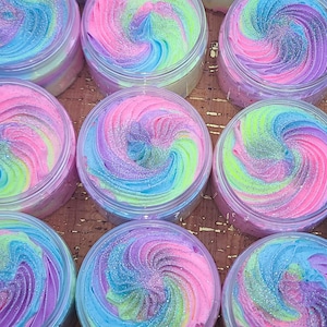 Puede incluir: Primer plano de ocho recipientes de jabón batido de colores. El jabón es un remolino de rosa, azul, verde y blanco con purpurina.