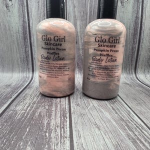 Op de afbeelding: Twee flessen Glo Girl Skincare Pumpkin Pecan Waffles Body Lotion. De flessen hebben een gemarmerd roze en grijs ontwerp met zwarte doppen. Elke fles is gelabeld met de merknaam, productnaam en ingrediënten.