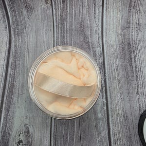 May include: A round, clear container with a peach-colored powder puff and a gold ribbon handle. The puff is soft and fluffy, and the container has a silver rim. The background is a wooden surface.