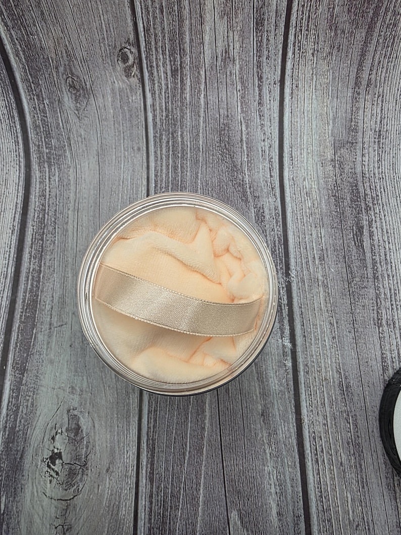 May include: A round, clear container with a peach-colored powder puff and a gold ribbon handle. The puff is soft and fluffy, and the container has a silver rim. The background is a wooden surface.