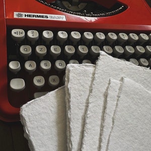 May include: A vintage red Hermes Baby typewriter with a white keyboard and a stack of white handmade paper on the right side.