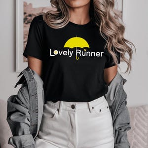 Puede incluir: Camiseta negra con un paraguas amarillo y el texto "Lovely Runner" impreso en la parte delantera.